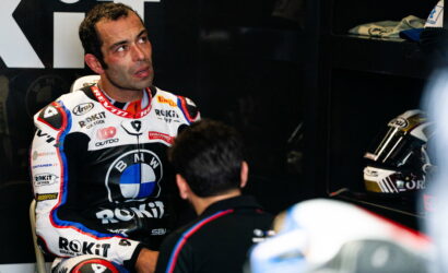 Motorcycle racer in sponsor-filled racing suit sits in a paddock, talking with a team member.