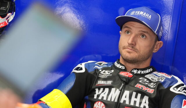 Motorcycle racer in Yamaha gear sitting against a blue wall, looking upward in thought.