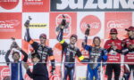 Five motorcycle racers on a podium hold up trophies, smiles on their faces, with a red-and-white sponsor banner backdrop behind them.