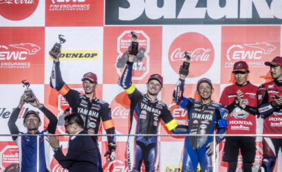 Five motorcycle racers on a podium hold up trophies, smiles on their faces, with a red-and-white sponsor banner backdrop behind them.