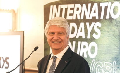 Senior man in a suit speaking at a podium with a 'INTERNATIONAL DAYS' banner behind him.