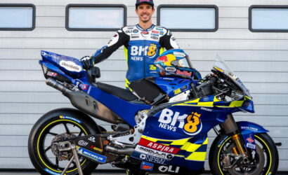 Rider in blue and yellow racing suit stands beside a blue Ducati race bike with sponsor stickers in front of a gray garage door.