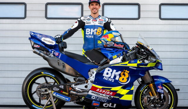Rider in blue and yellow racing suit stands beside a blue Ducati race bike with sponsor stickers in front of a gray garage door.