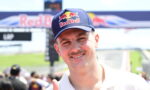 Man wearing a navy Red Bull cap smiles at a sunny race track with a finish line in the background, giving a thumbs-up.