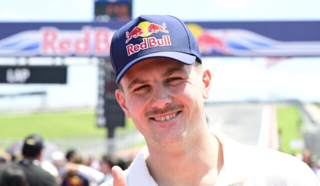Man wearing a navy Red Bull cap smiles at a sunny race track with a finish line in the background, giving a thumbs-up.