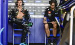 Two motorcycle racers in a pit area: left rider in open black leather suit with headset, right team member writing on a clipboard against sponsor banners.