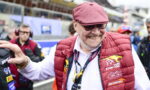 Smiling older man wearing a pink-red flat cap and a red padded vest at a race track, posing for the camera.
