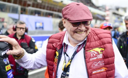 Smiling older man wearing a pink-red flat cap and a red padded vest at a race track, posing for the camera.
