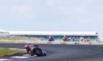 MotoGP rider in red and blue leans into a left-hand turn on a race track, with other riders in the background.