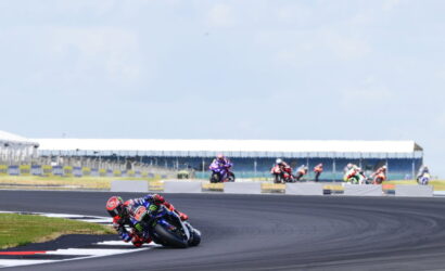 MotoGP rider in red and blue leans into a left-hand turn on a race track, with other riders in the background.