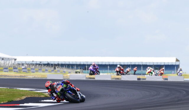MotoGP rider in red and blue leans into a left-hand turn on a race track, with other riders in the background.