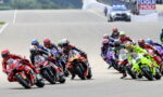 Group of motorcycle racers leaning into a turn on a sunny track during a race in close formation.