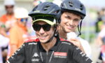 Two cyclists wearing helmets and race jerseys pose for a photo, smiling at the camera, front rider with sunglasses and neon-green visor cap.