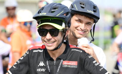 Two cyclists wearing helmets and race jerseys pose for a photo, smiling at the camera, front rider with sunglasses and neon-green visor cap.