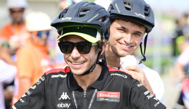 Two cyclists wearing helmets and race jerseys pose for a photo, smiling at the camera, front rider with sunglasses and neon-green visor cap.