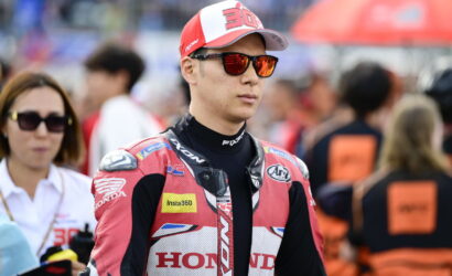 MotoGP rider in a red Honda racing suit and cap with the number 30, wearing orange sunglasses in the paddock setting