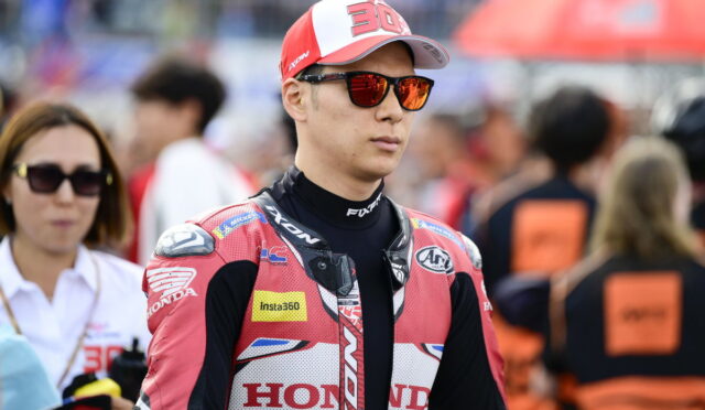 MotoGP rider in a red Honda racing suit and cap with the number 30, wearing orange sunglasses in the paddock setting