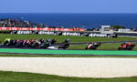 Motorcycle race on a coastal track with riders leaning into a left-hand turn; sea and stands in the background, banners along the barrier.