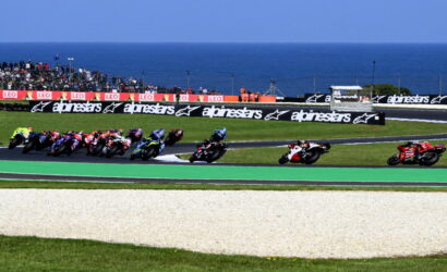 Motorcycle race on a coastal track with riders leaning into a left-hand turn; sea and stands in the background, banners along the barrier.