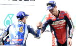 Two motorcycle racers shake hands on a podium, one in blue gear with CAT and Michelin logos and the other in red Castrol/Honda gear.