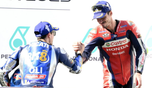Two motorcycle racers shake hands on a podium, one in blue gear with CAT and Michelin logos and the other in red Castrol/Honda gear.