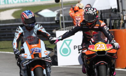 Two motorcycle racers on a race track give a high-five as they ride side by side; one in light-blue/orange Aprilia, the other in a Red Bull KTM #23 livery.