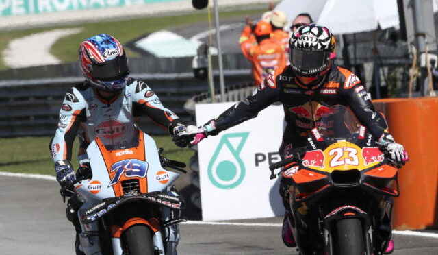 Two motorcycle racers on a race track give a high-five as they ride side by side; one in light-blue/orange Aprilia, the other in a Red Bull KTM #23 livery.