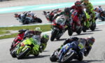 Group of motorcycle racers leaning into a tight turn on a racetrack, neon green bike leading the pack and blue bike close beside.