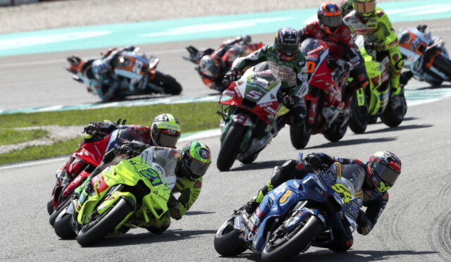 Group of motorcycle racers leaning into a tight turn on a racetrack, neon green bike leading the pack and blue bike close beside.