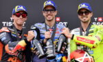 Three motorcycle racers on a podium in branded suits and blue Michelin caps, toasting with Prosecco bottles.