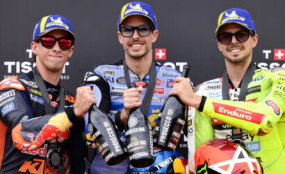 Three motorcycle racers on a podium in branded suits and blue Michelin caps, toasting with Prosecco bottles.