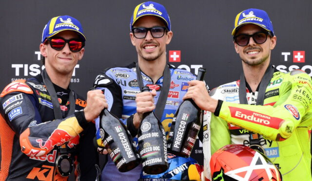 Three motorcycle racers on a podium in branded suits and blue Michelin caps, toasting with Prosecco bottles.