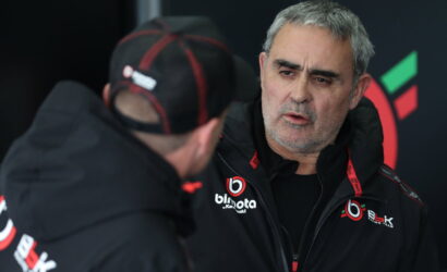 Two men in black jackets with team logos talking in a pit area; one faces the camera with a gray beard.