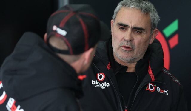 Two men in black jackets with team logos talking in a pit area; one faces the camera with a gray beard.