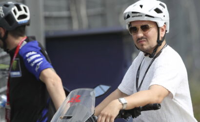 Man wearing a white bike helmet and sunglasses sits on a scooter or bike at a racetrack, with a teammate blurred in the background.
