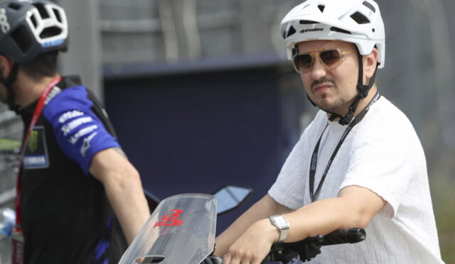 Man wearing a white bike helmet and sunglasses sits on a scooter or bike at a racetrack, with a teammate blurred in the background.