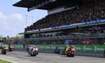 Motorcycle race on a track with a packed grandstand; riders in colorful gear speed off from the start as fans cheer.