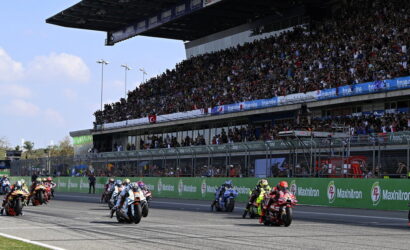 Motorcycle race on a track with a packed grandstand; riders in colorful gear speed off from the start as fans cheer.