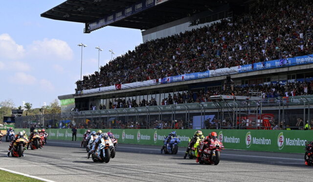 Motorcycle race on a track with a packed grandstand; riders in colorful gear speed off from the start as fans cheer.