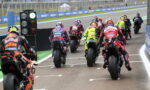 Motorcycle racers in colorful racing suits lined up at the starting grid on a race track with a green starting light visible on the left.