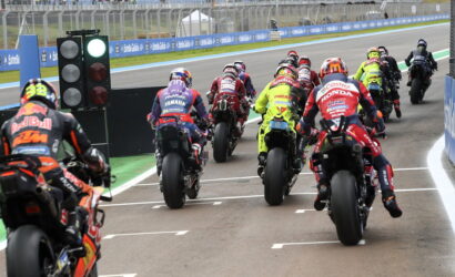 Motorcycle racers in colorful racing suits lined up at the starting grid on a race track with a green starting light visible on the left.