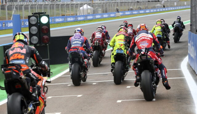 Motorcycle racers in colorful racing suits lined up at the starting grid on a race track with a green starting light visible on the left.