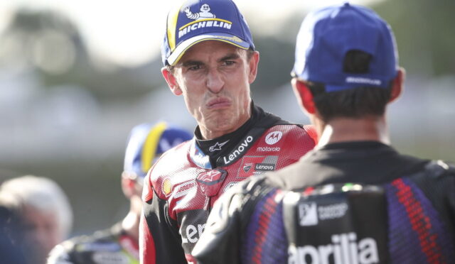 MotoGP rider in red Lenovo suit making a funny face toward another rider wearing a blue cap.
