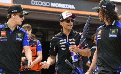 Three race team members in blue-black uniforms chat together, sponsor logos visible in a pit area backdrop.