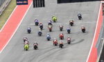 Group of colorful motorcycle racers blasting off from the start on a race track, Red Bull banner overhead.