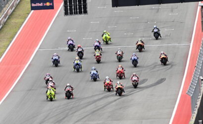 Group of colorful motorcycle racers blasting off from the start on a race track, Red Bull banner overhead.