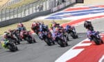 Group of sport motorcycles racing into a left-hand turn on a race track, riders leaning at speed; red and white curbs visible in the foreground.