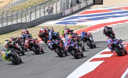 Group of sport motorcycles racing into a left-hand turn on a race track, riders leaning at speed; red and white curbs visible in the foreground.