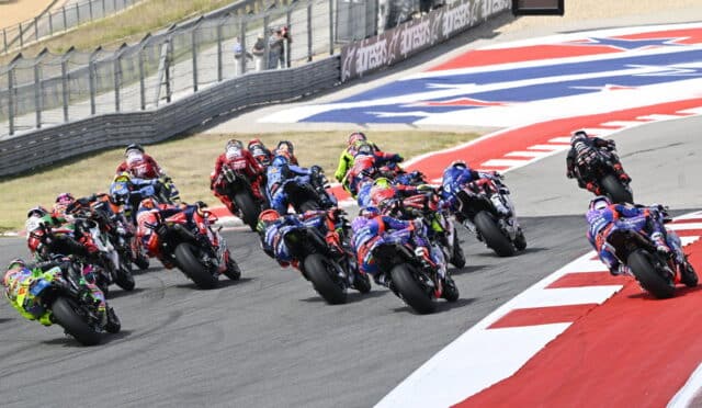 Group of sport motorcycles racing into a left-hand turn on a race track, riders leaning at speed; red and white curbs visible in the foreground.
