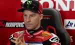 Motorcycle racer in a red Honda suit sits in a chair during an interview, wearing a Monster cap and sunglasses on top of the cap.